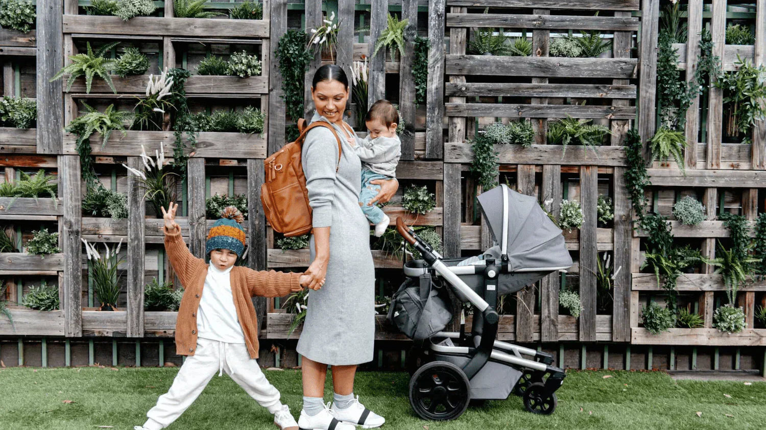 Woman carrying a baby and a nappy bag filled with essentials, holding her toddler with a pram beside her.