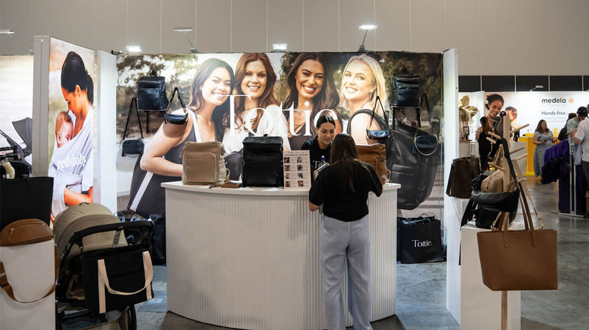 A woman stands in front of a Tottie booth at the One Fine Baby Expo, showcasing a variety of stylish handbags and nappy bags