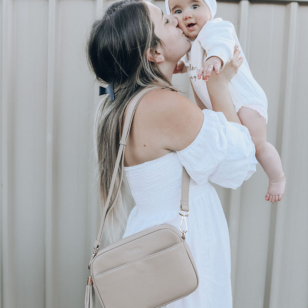 a mother carrying and kissing her baby and carrying her stone colored leather crossbody bag