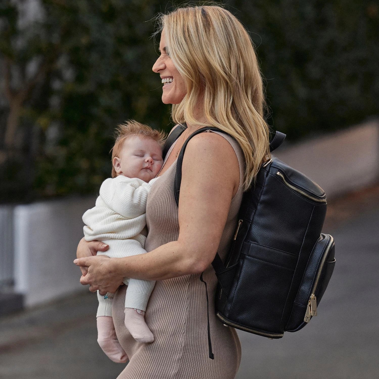 Mother holding her child with a stylish black nappy bag backpack featuring gold hardware.