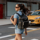 Stylish woman wearing a denim romper carrying a black backpack with gold hardware while walking on a city street.
