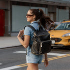 Woman wearing a denim romper walking down the street with a black backpack featuring gold hardware.