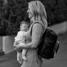 Black and white photo of a woman carrying her child while wearing a black nappy bag backpack