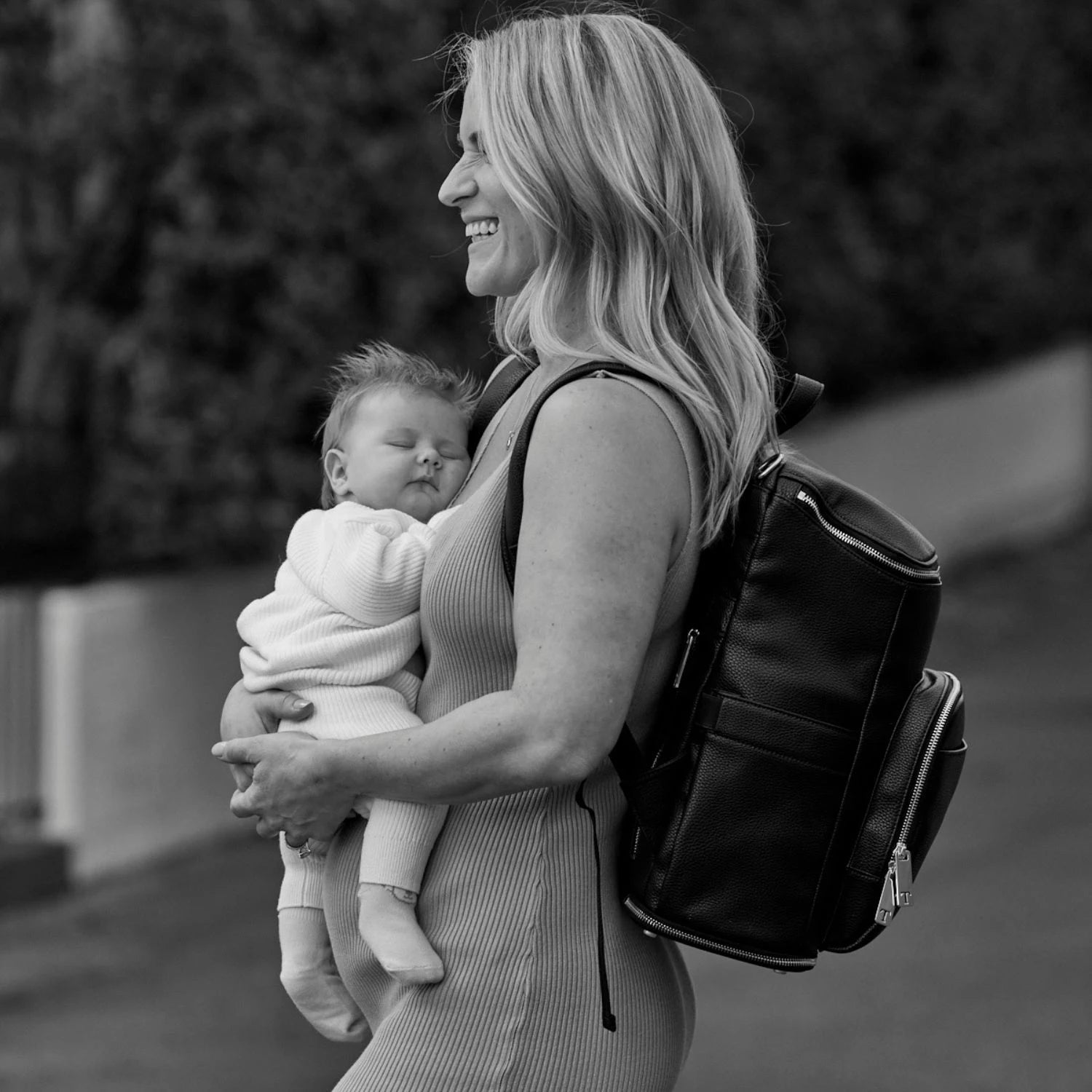 Black and white photo of a woman carrying her child while wearing a black nappy bag backpack