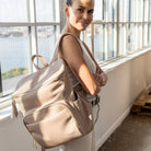 Woman seen from the side carrying a stone-colour nappy bag backpack accented with gold hardware on her shoulder.