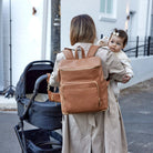 Back view of a woman carrying her baby and pushing a baby pram while wearing a tan nappy bag backpack with gold hardware.