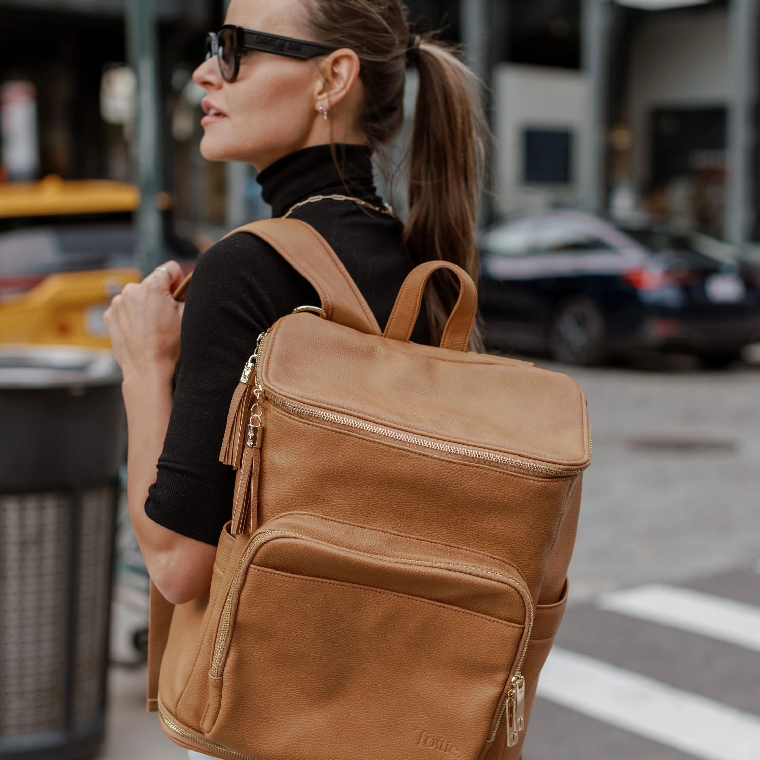 Side view of a woman wearing a black top and sunglasses carrying a tan nappy bag backpack with gold hardware.