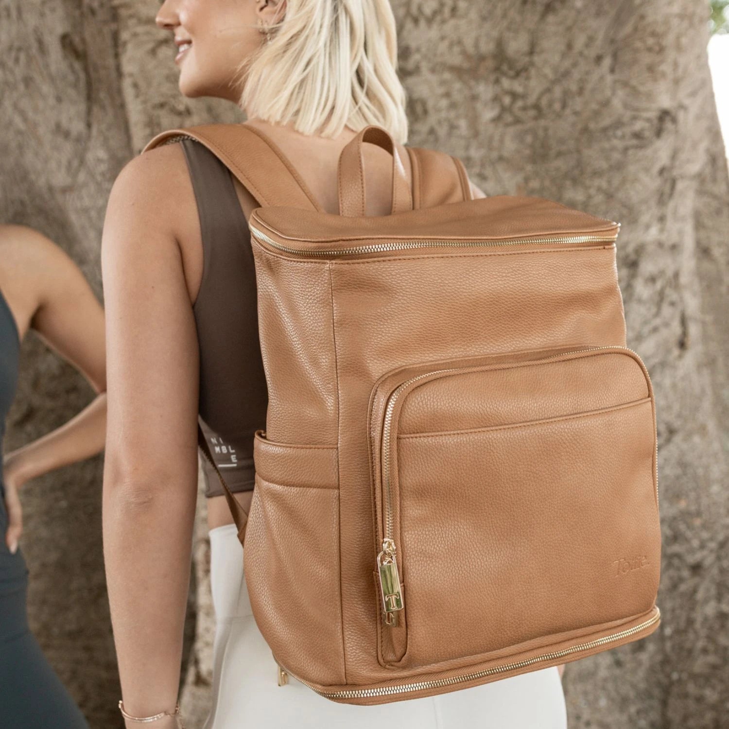 Back profile of a woman in a summer outfit with a tan nappy bag backpack featuring gold hardware