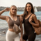 Two women wearing summer outfits; one carrying a stone-colour bum bag, the other with a tan nappy bag backpack featuring gold hardware.