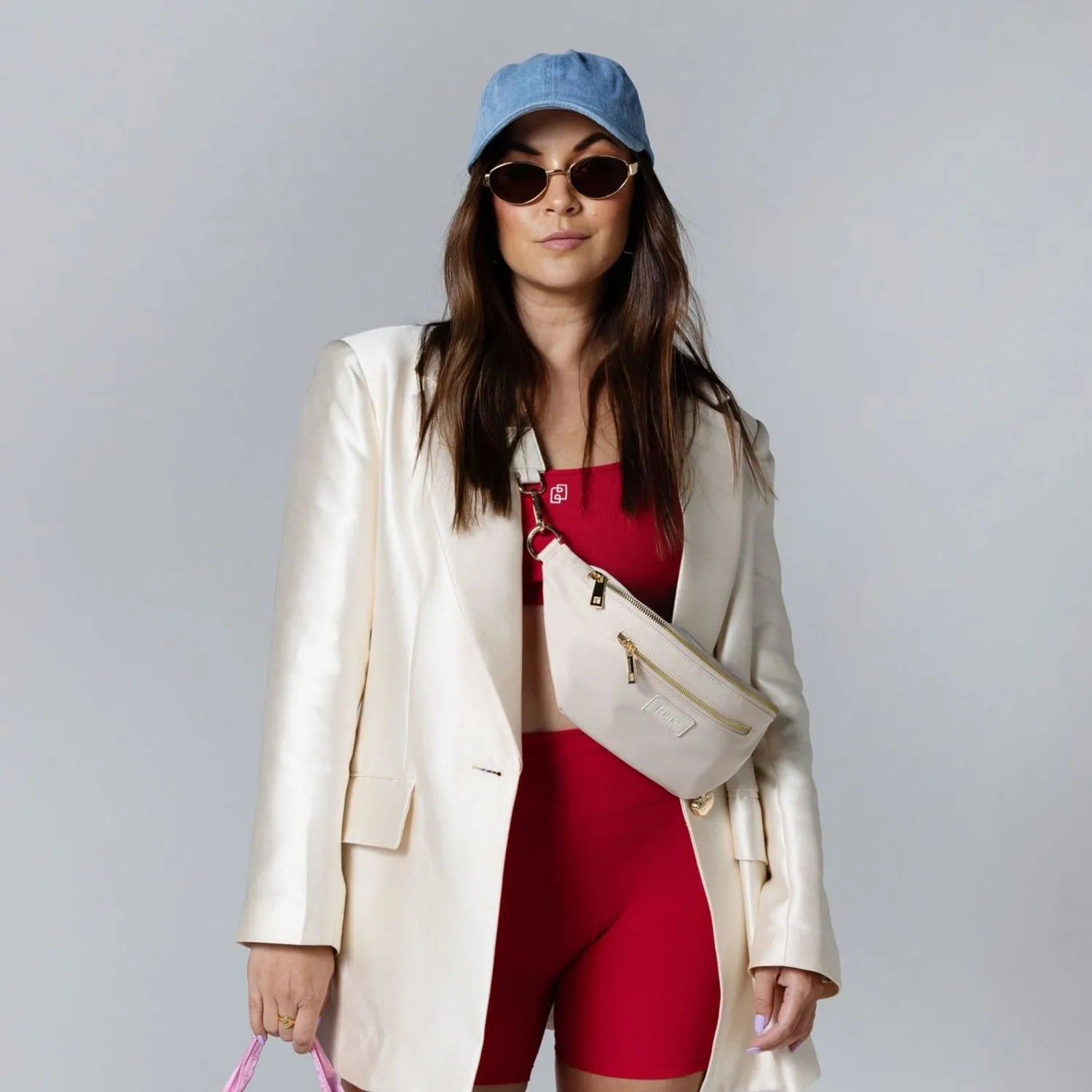 Stylish woman in blue outfit and cream coat carrying cream nylon bum bag, wearing sunglasses and blue cap.