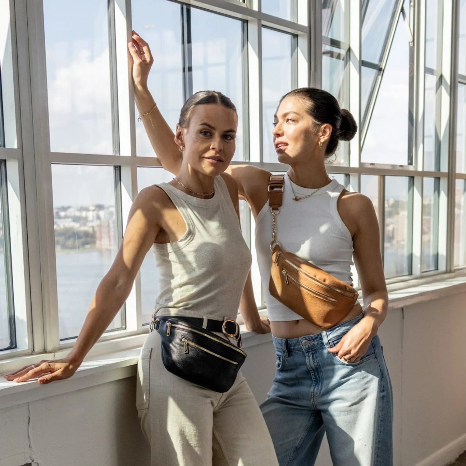 Two women side by side wearing black bum bag with gold hardware and tan bum bag with gold hardware.