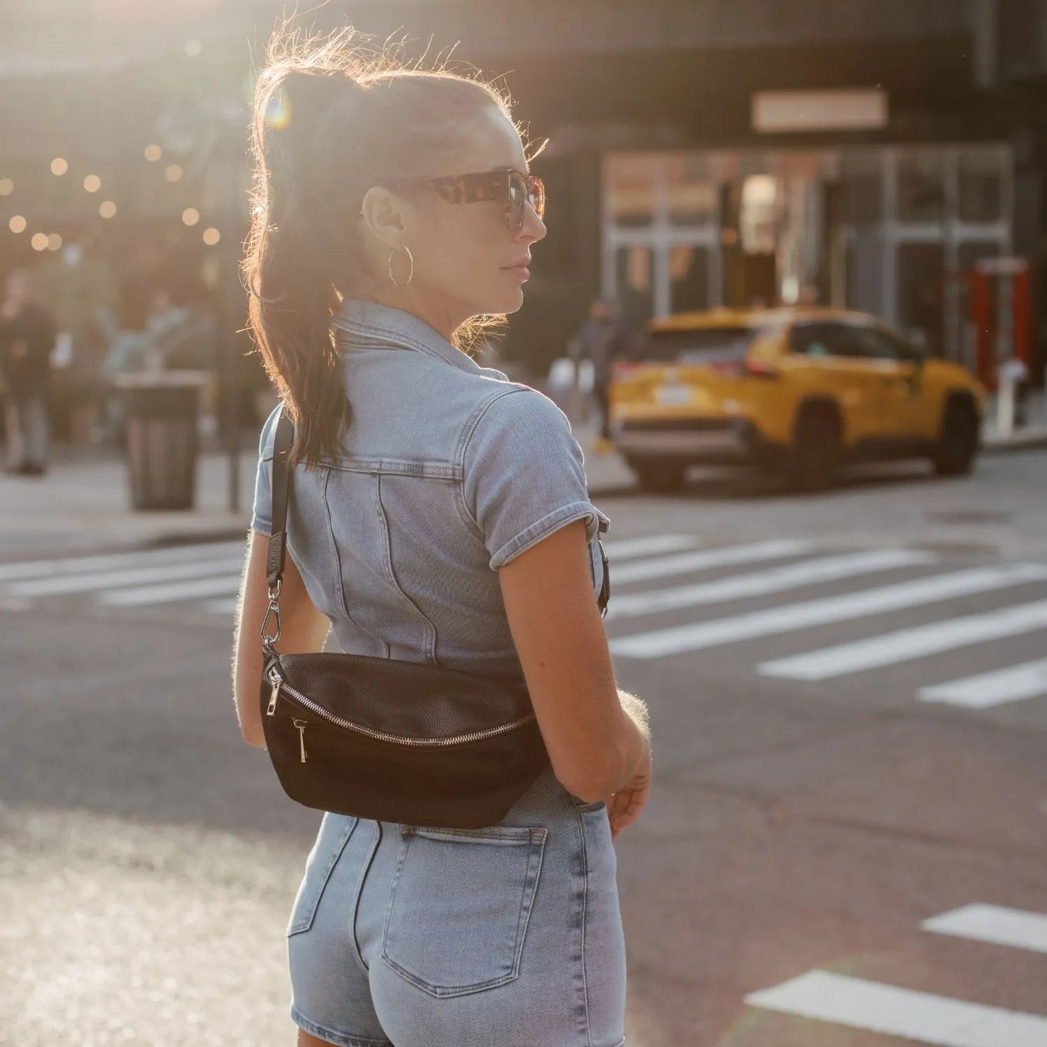 Woman wearing a black bum bag with silver hardware crossbody style on her back, walking on a blurred street background.