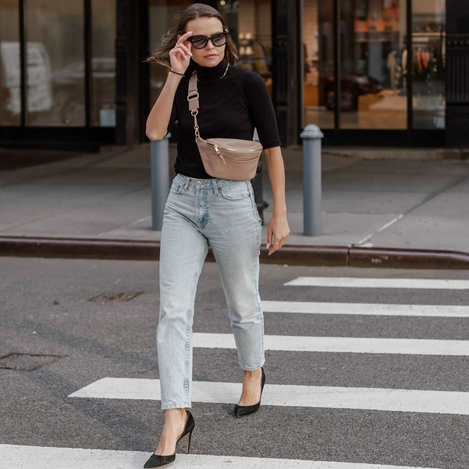 Woman crossing a walking street wearing a stone colour bum bag with gold hardware in front crossbody style, styled with black top and skinny jeans