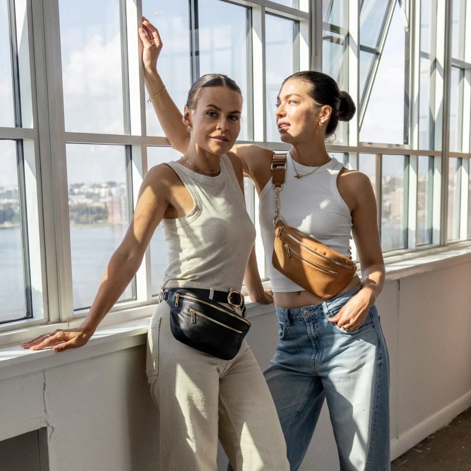 Two women wearing bum bags – one styles a black waist bag with gold hardware, the other wears a tan crossbody bum bag