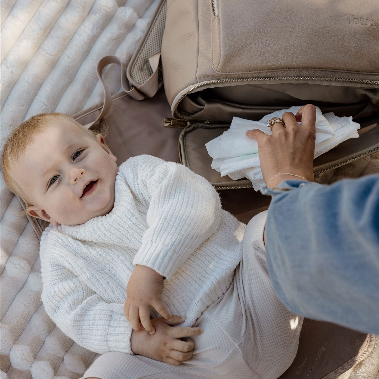 Baby lying on a padded baby change mat next to an open beige nappy bag with tissues, wearing a white knit outfit. Mum reaching for piece of nappy, ready for nappy changing