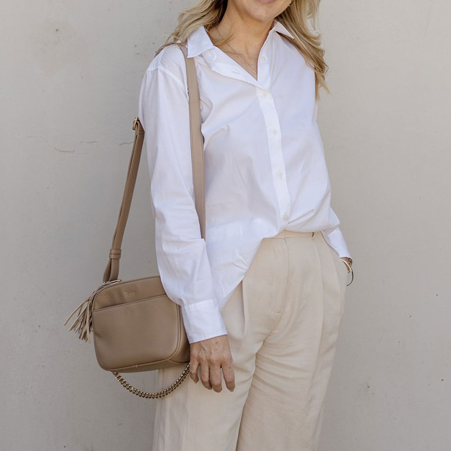 A woman wearing a white shirt and beige pants holding a beige handbag against a plain background