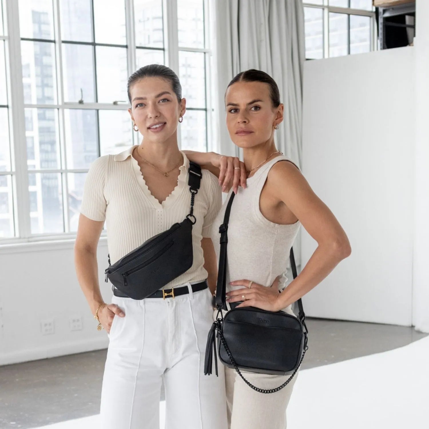 Two women dressed in white, carrying black crossbody and bum bags with black hardware.