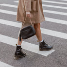 Woman wearing brown coat and boots carrying black crossbody bag with gold hardware while crossing pedestrian.