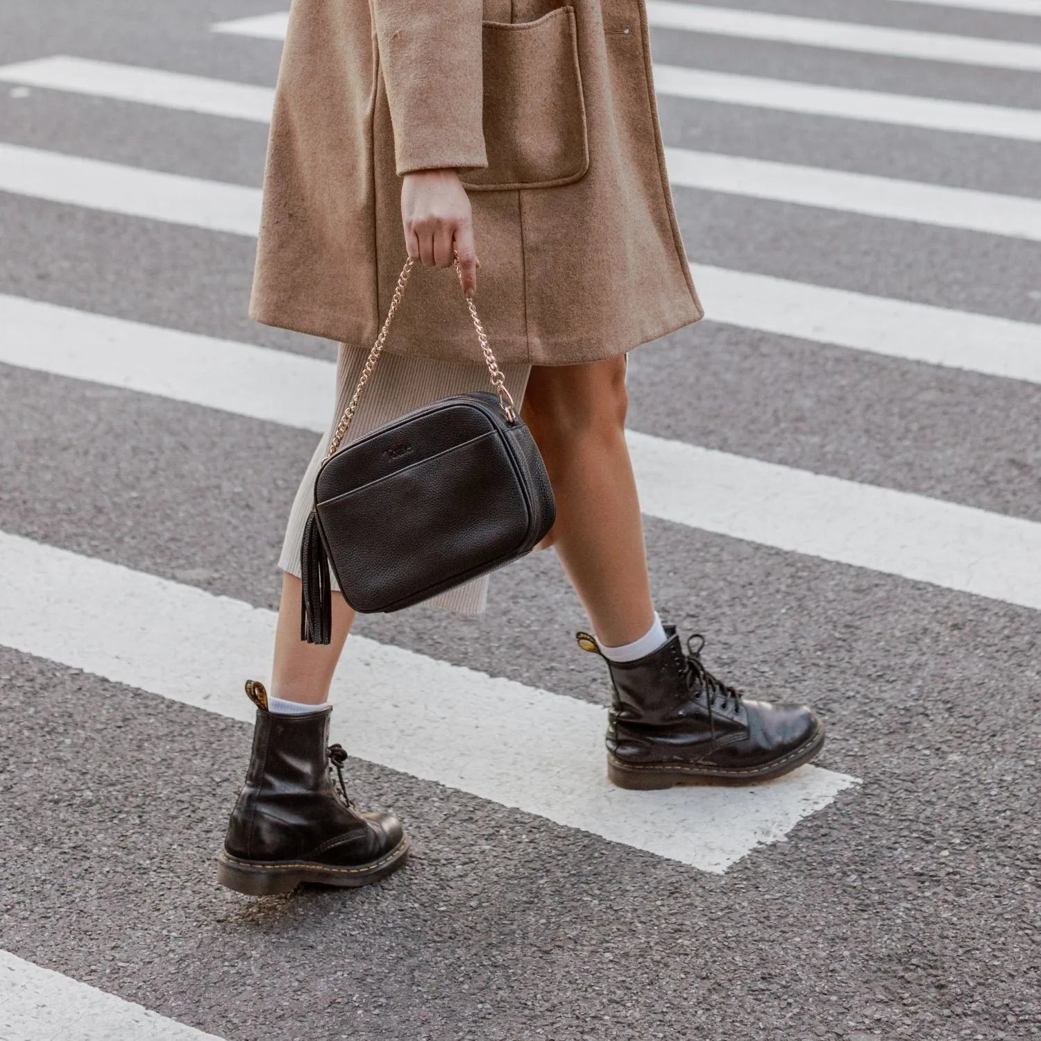 Woman wearing brown coat and boots carrying black crossbody bag with gold hardware while crossing pedestrian.