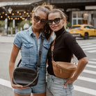 Woman carrying black crossbody bag with silver hardware beside another wearing tan bum bag with gold hardware.