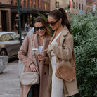 Two women in coats, one carrying tan crossbody bag with gold hardware and showing phone, the other with stone crossbody bag on shoulder.