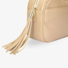 Detailed view of gold hardware tassel on stone colour crossbody bag on white background.