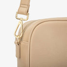 Close-up of gold hardware detail with stone colour strap on stone colour crossbody bag against white background.