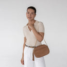 Casual woman posing with tan colour crossbody bag featuring gold hardware.