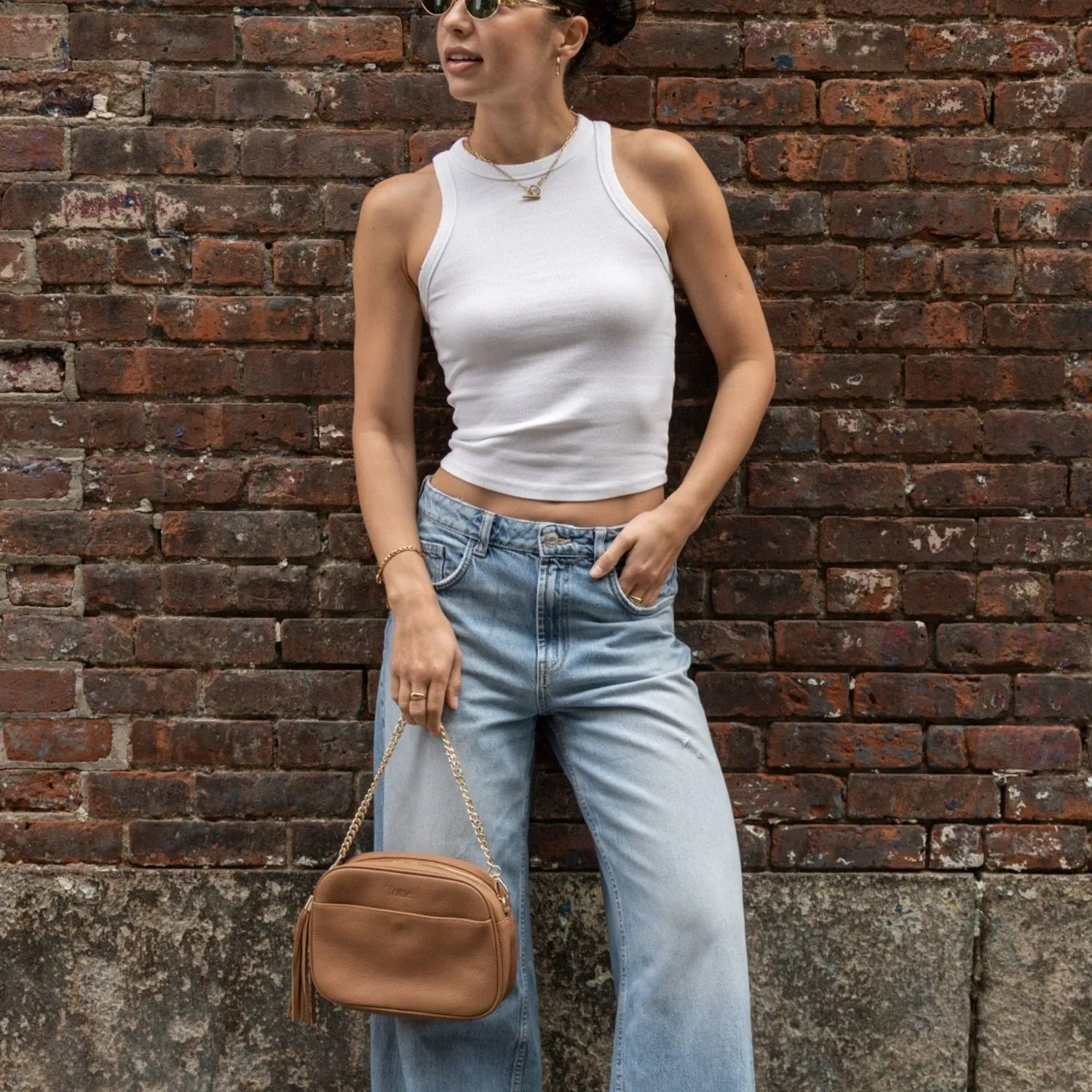 Stylish woman in sunglasses holding tan colour crossbody bag featuring gold hardware.