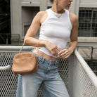 Tan colour crossbody bag with gold hardware carried by woman in white top and jeans.