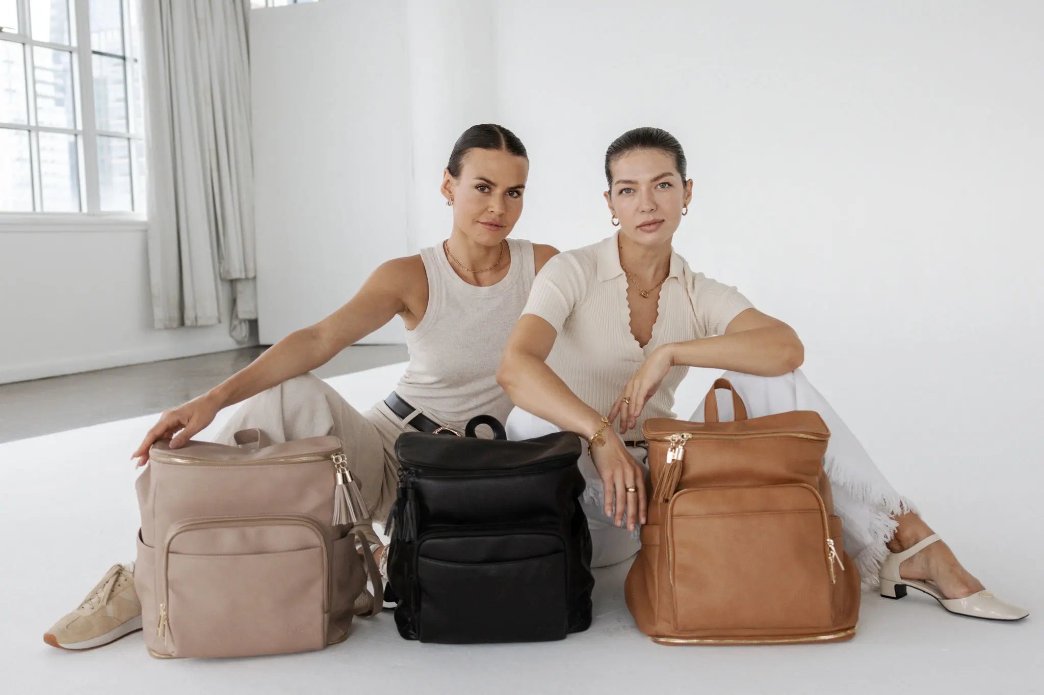 Three stylish nappy bags in stone, tan, and black colours shown by two women sitting.