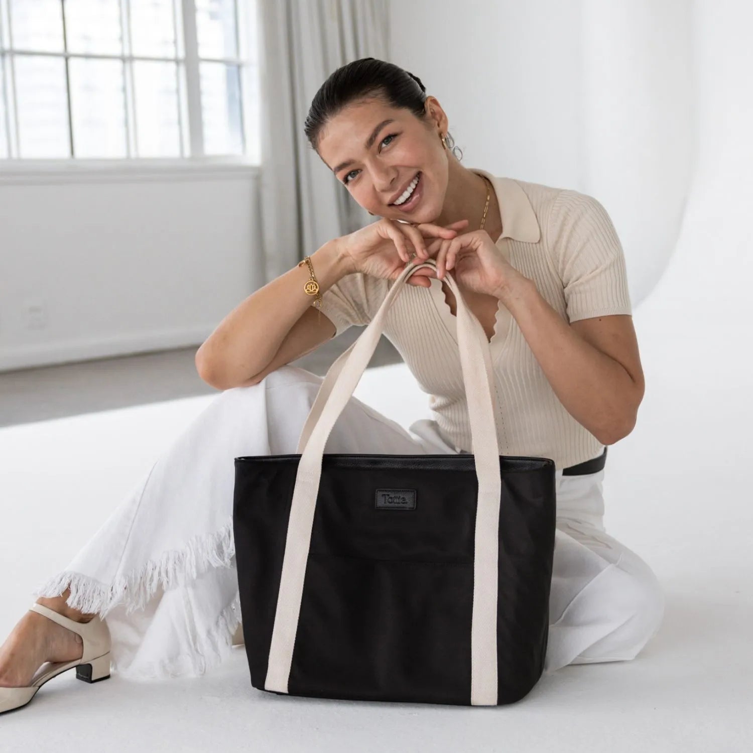 Black nylon tote bag with cream strap close-up, carried by woman in cream and white outfit.
