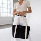 Detailed view of black nylon tote bag with cream strap, woman dressed in cream top and white pants.