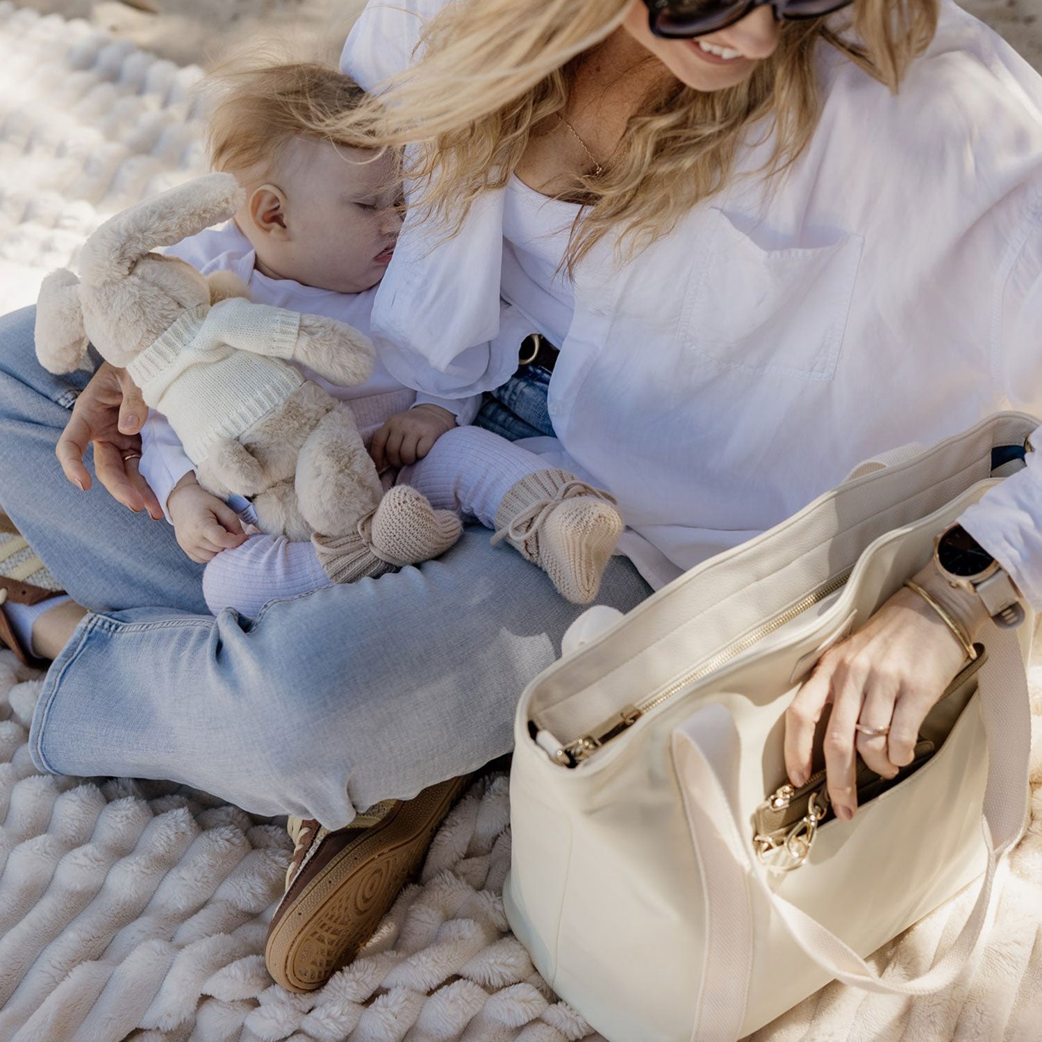 Woman sitting on a beach mat reaching for her card holder wallet inside her nylon tote bag in yellow, while her baby lying on her lap holding a teddy bear, both wearing white outfits.