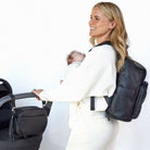 Woman carrying a baby and holding a black stroller. The stroller has a black crossbody bag from Tottie attached to the black pram clips. The woman is also wearing a black nappy backpack from Tottie