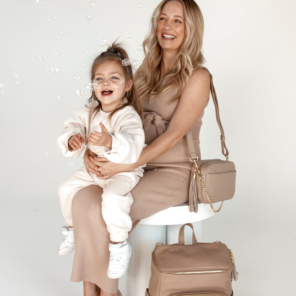 a pregnant woman holding her child and carrying a bestie crossbody bag with a baby backpack beside her