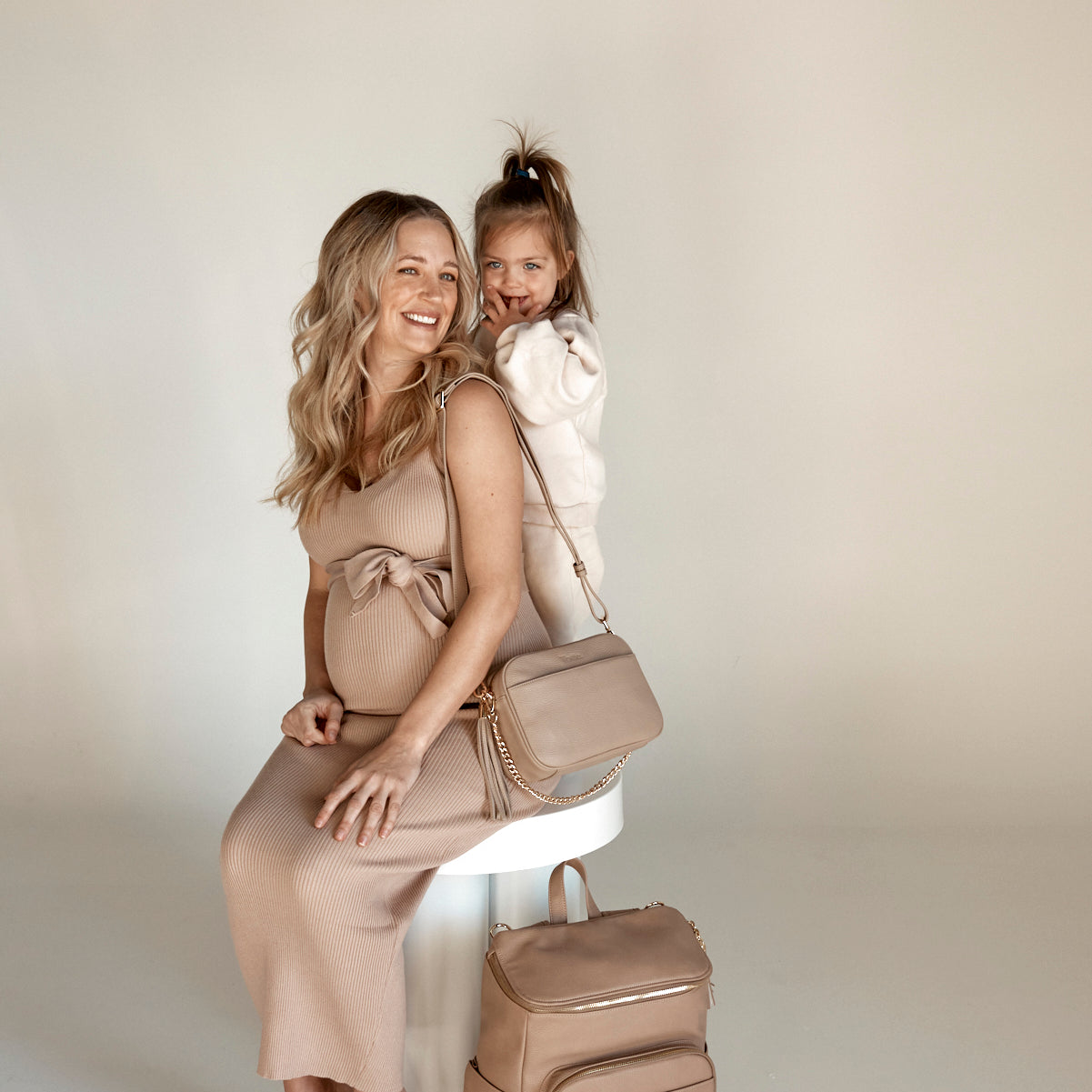 a pregnant woman and a child on her back while carrying a stone bestie crossbody bag and stone nappy bag backpack from Tottie