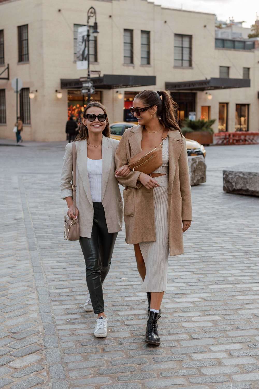 Two women stroll down a brick street, wearing women's bags from Tottie. One is wearing a stone crossbody bag, and the other is wearing a tan bum bag.