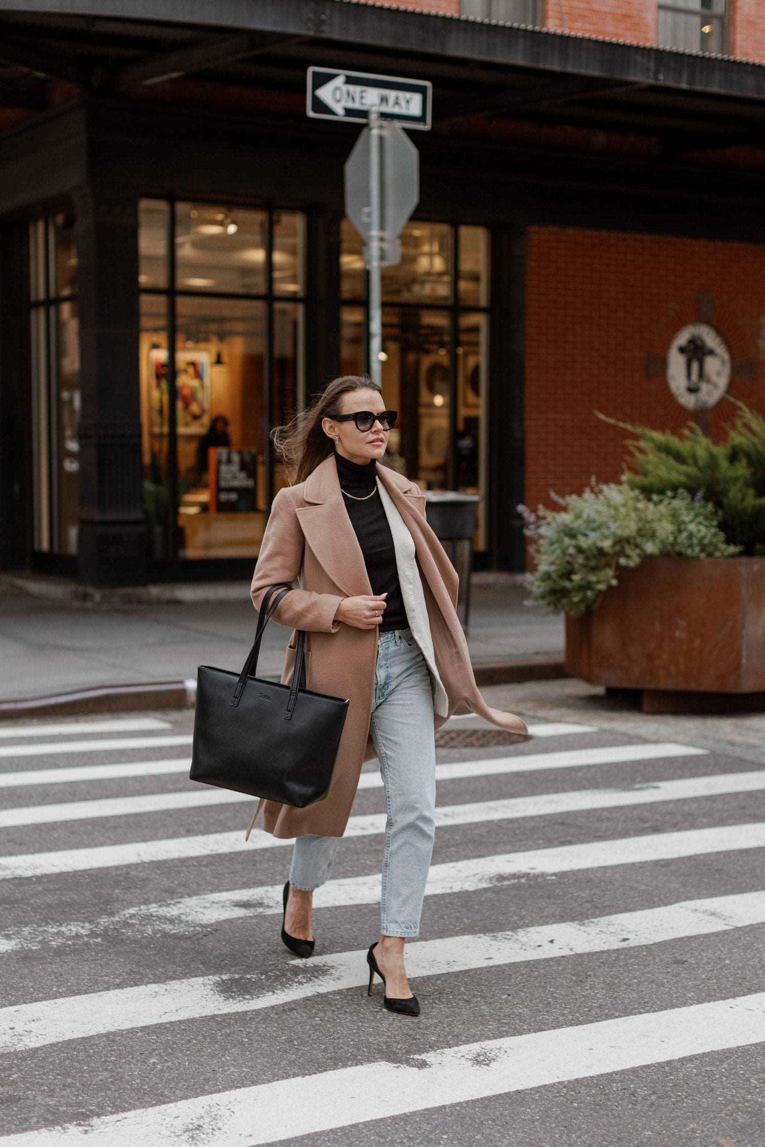 A woman in a coat and heels crosses the street while carrying a black tote bag