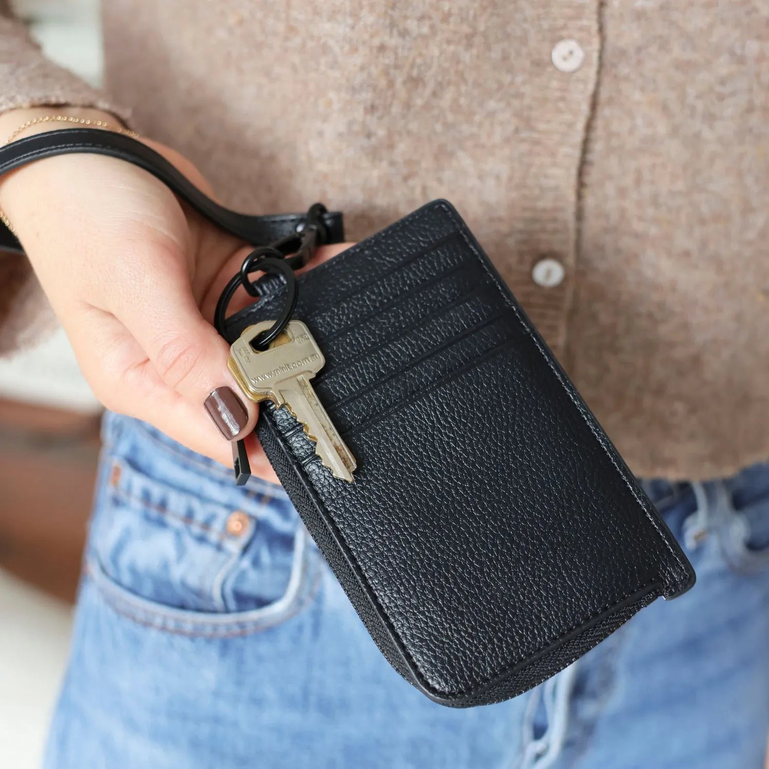 Hand holding a black card holder wallet wristlet with a key, against a blurred background of jeans and a brown top."