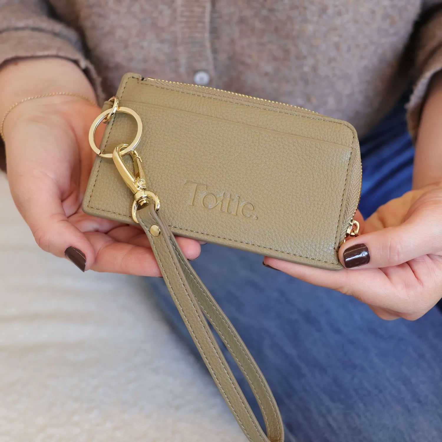 Stylish khaki card holder wallet wristlet with key, held by hand with blurred jeans and brown top background.