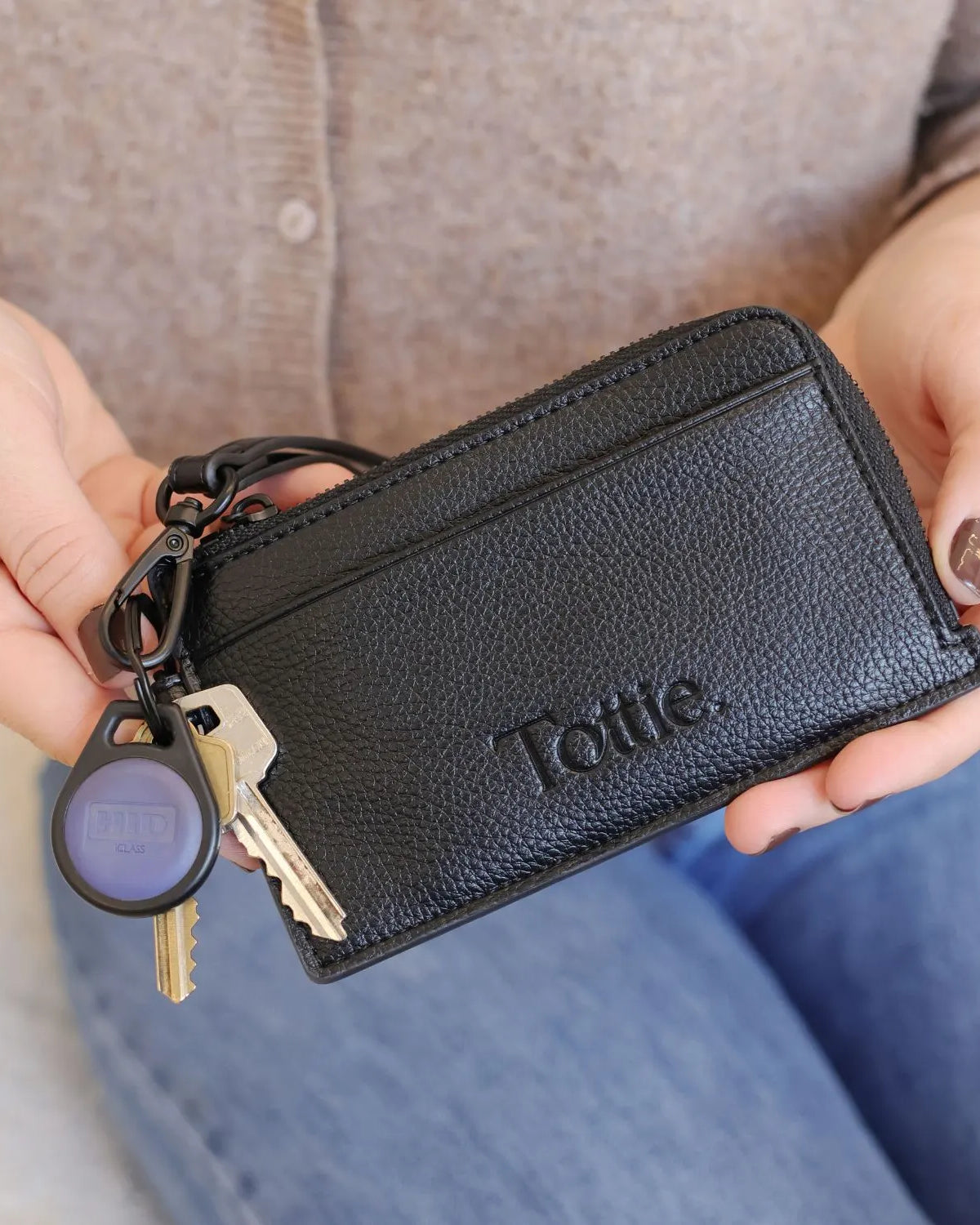 Black card holder wristlet wallet held in front of blurred denim jeans and brown top.