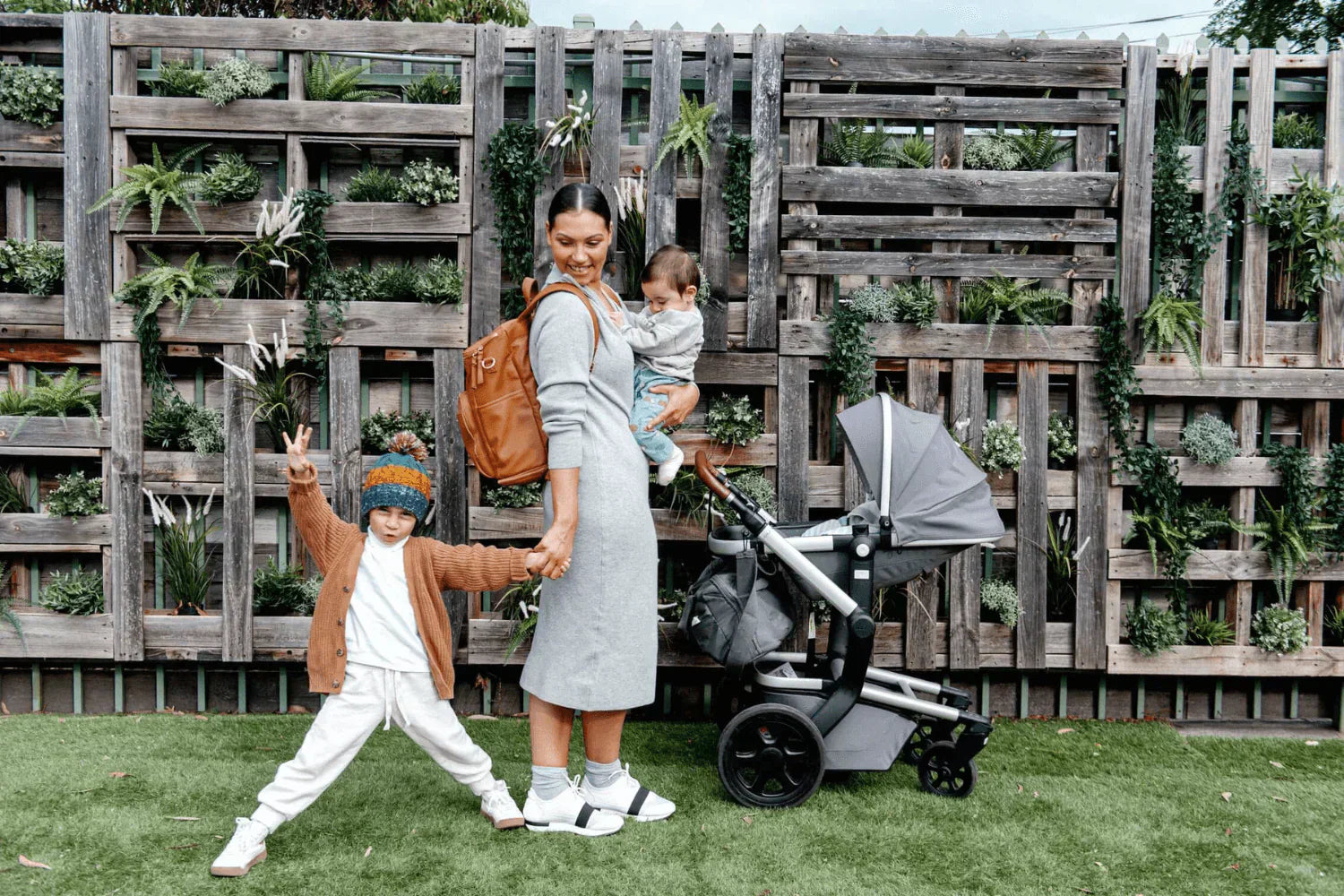 Woman carrying a baby and a nappy bag filled with essentials, holding her toddler with a pram beside her.