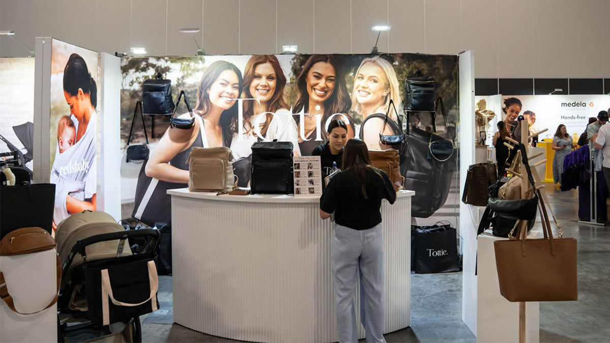 A woman stands in front of a Tottie booth at the One Fine Baby Expo, showcasing a variety of stylish handbags and nappy bags