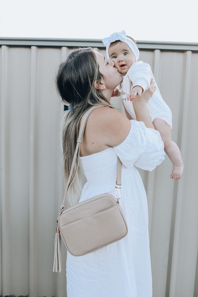 a mother carrying and kissing her baby and carrying her stone colored leather crossbody bag