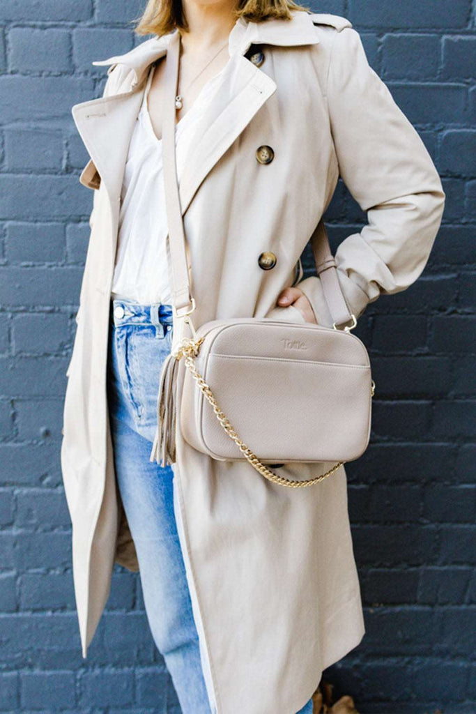 woman wearing a trench coat holding and carrying a stone colored leather crossbody bag
