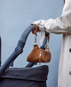 A woman pushing a stroller, with a tan colour bag with pram clips in gold hanging on the stroller