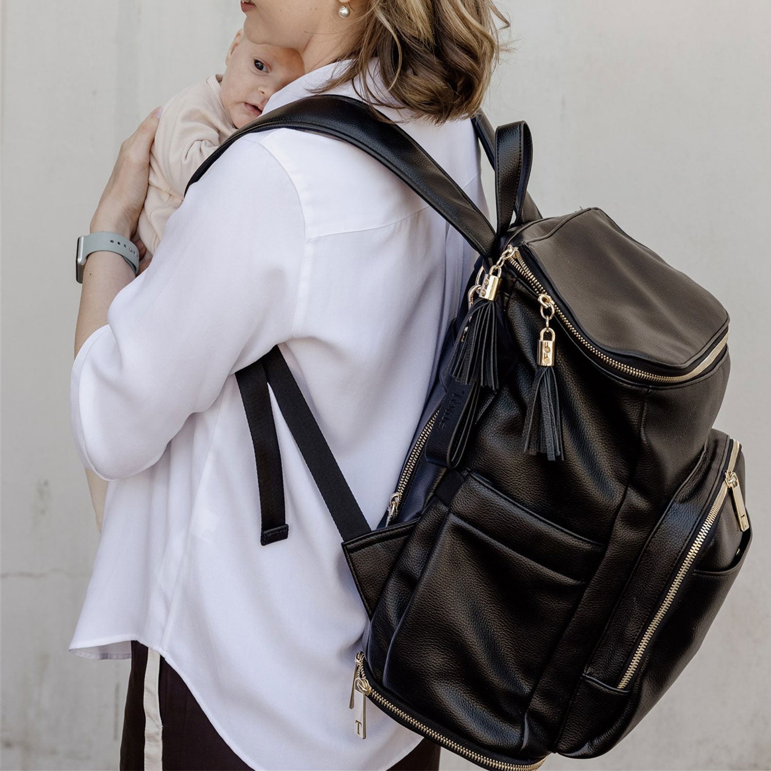 Person wearing a black nappy bag backpack with gold zippers against a plain background