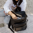 Person sitting on a sandy beach with a black baby bag backpack and a baby breastfeeding