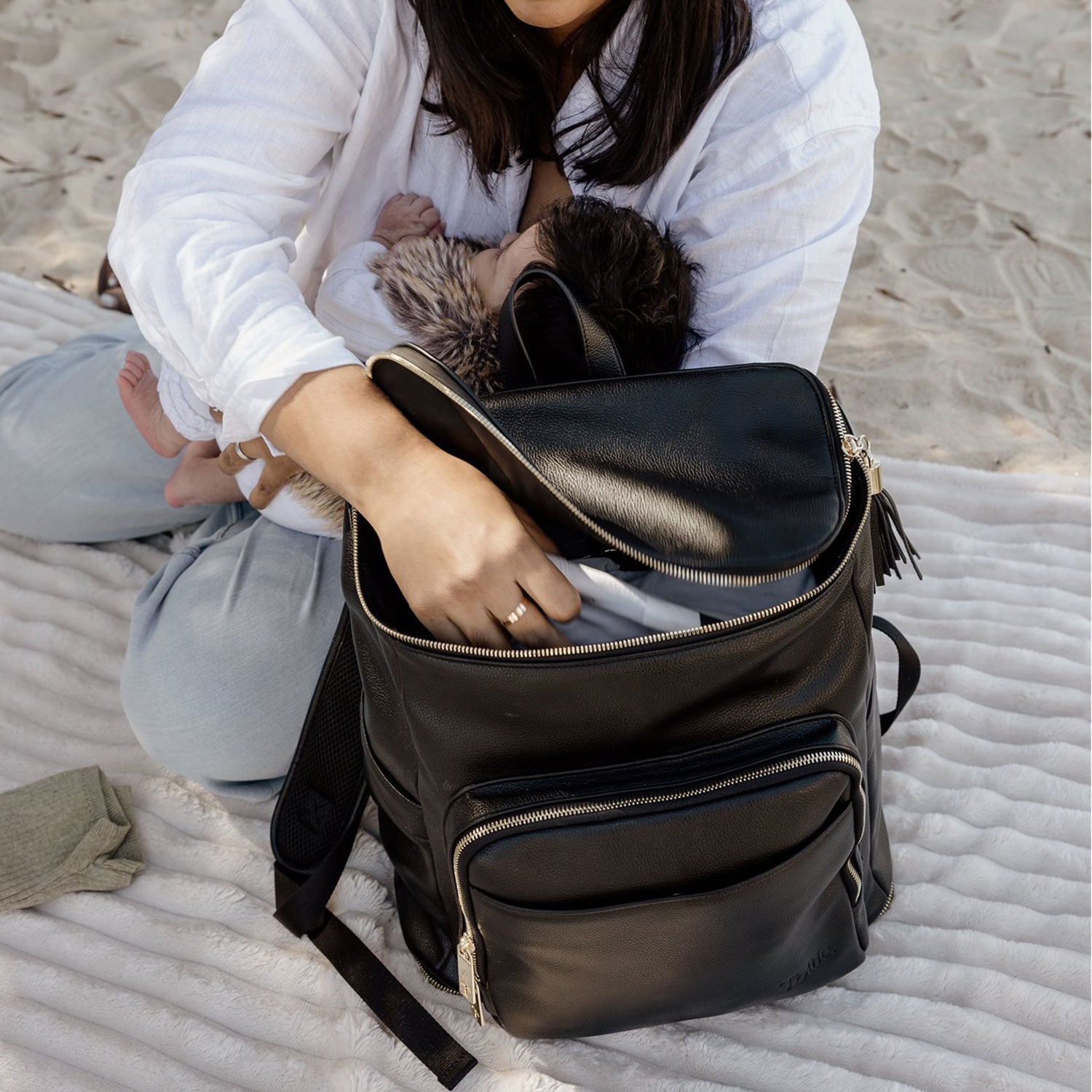 Person sitting on a sandy beach with a black baby bag backpack and a baby breastfeeding
